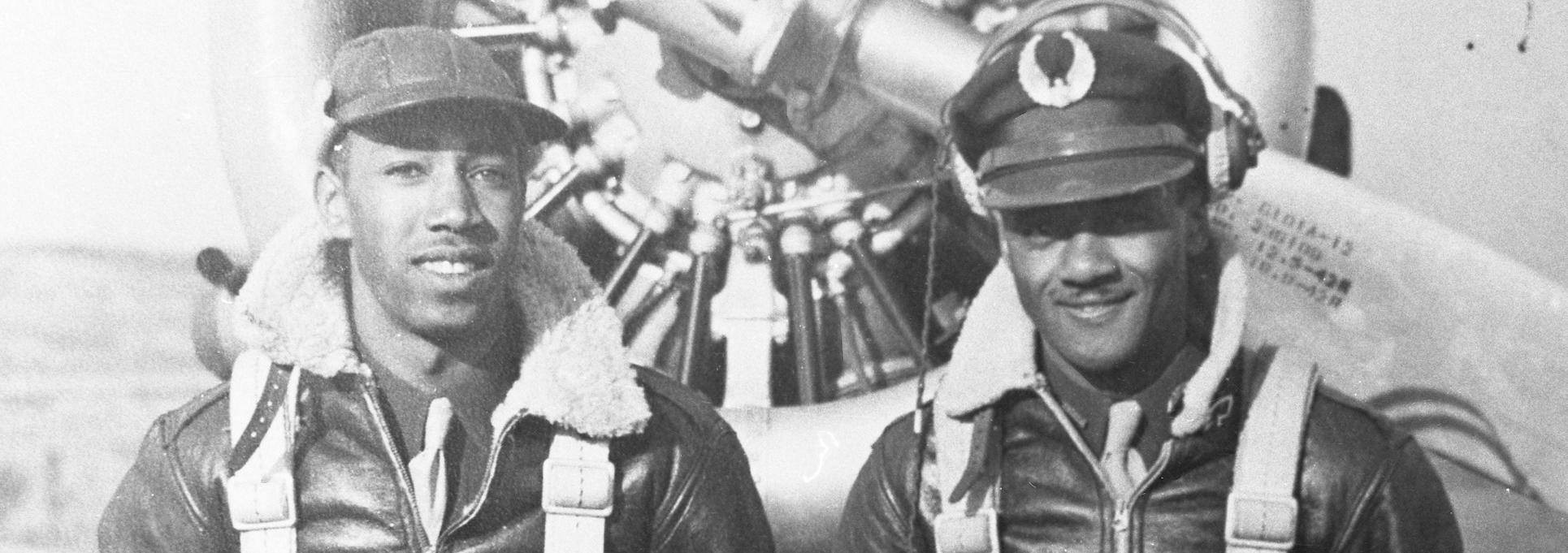 Black and white photo with two African American men wearing bomber jackets and harnesses standing in front of a propeller plane