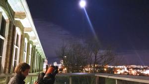 Woman stands makes adjustment to telescope pointed at Moon which is visible in the night sky from the Observatory Terrace. Columbia skyline visible in background.