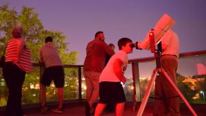Group of people stand on terrace in the early evening with one young boy looking at the night sky through binoculars in the foreground