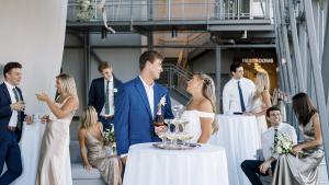 Bride and groom stand behind small, tall table with white tablecloth and members of their wedding party socializing in the background with the side of the planetarium dome exterior visible on the left side of the frame.