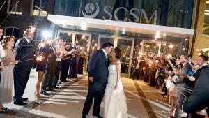 Bride and groom pose at museum entrance at night with guests holding sparklers along either side of the couple