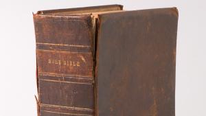Large brown leather bound Bible stand vertically with spine facing camera and "Holy Bible" written on it.