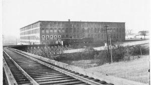 Black and white photograph shows train tracks in the foreground with a large brick, four story building in the background