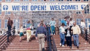 Group of people walking up stairs towards a large brick building with a banner over the entrance that reads &amp;quot;We're Open! Welcome!&amp;quot;