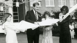 Black and white images shows 4 adults (1 man and 3 women) cutting through a large ribbon with an oversize pair of scissors.