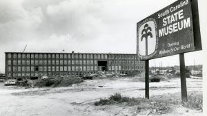 Black and white image of long, four story textile mill building under renovation. A sign is posted in front that reads &amp;quot;South Carolina State Museum Opening Windows to Our World&amp;quot;.