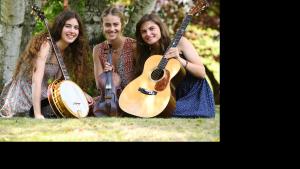 Three young women kneel on the grass. On the left the woman has long dark hair and holds a banjo, inter the center the woman has her hair tied back and holds a fiddle, and the women on the right also has long dark hair and she holds a guitar.