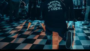 Dark room with bright spotlights and checkered dance floor shows line of men clapping for a dance kneeling on the floor with his back to camera wearing a dark colored hoodie