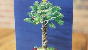 Christmas Card Depicts Illustrated Palmetto Tree Wrapped in Holiday Lights With Presents at the Base