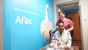 Woman pushes a wheelchair with another woman sitting in it through a space with a large blue poster featuring a duck and name Aflac on the left side and a large, changing table on the right side.