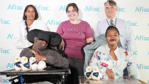 Three adults stand in front of a photo back drop that reads &amp;quot;Aflac&amp;quot; with two adults in wheelchairs in front of them.