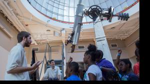 Bearded man on left side of the image addresses a group of young students with a huge telescope just behind them.