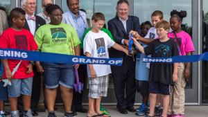 Group of older and younger people stand behind a blue ribbon that is being cut by a man holding an oversize pair of blue handled scissors.