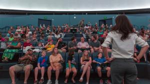 Woman with long dark hair stands with her back to the camera. An audience of people is seated in front of her in rows of blue chairs under a large, domed projector screen.