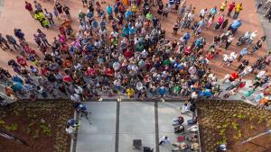 Image looks down from a large height onto a large group of people standing below, in front of the main entrance to a building.