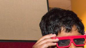 Young boy in yellow t-shirt with hands up to either side of his eyes as he adjusts a pair of red 3D glasses