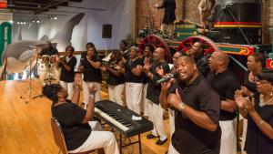 Group of singers, keyboard player and drummer in black shirts perform in front of a life size train replica