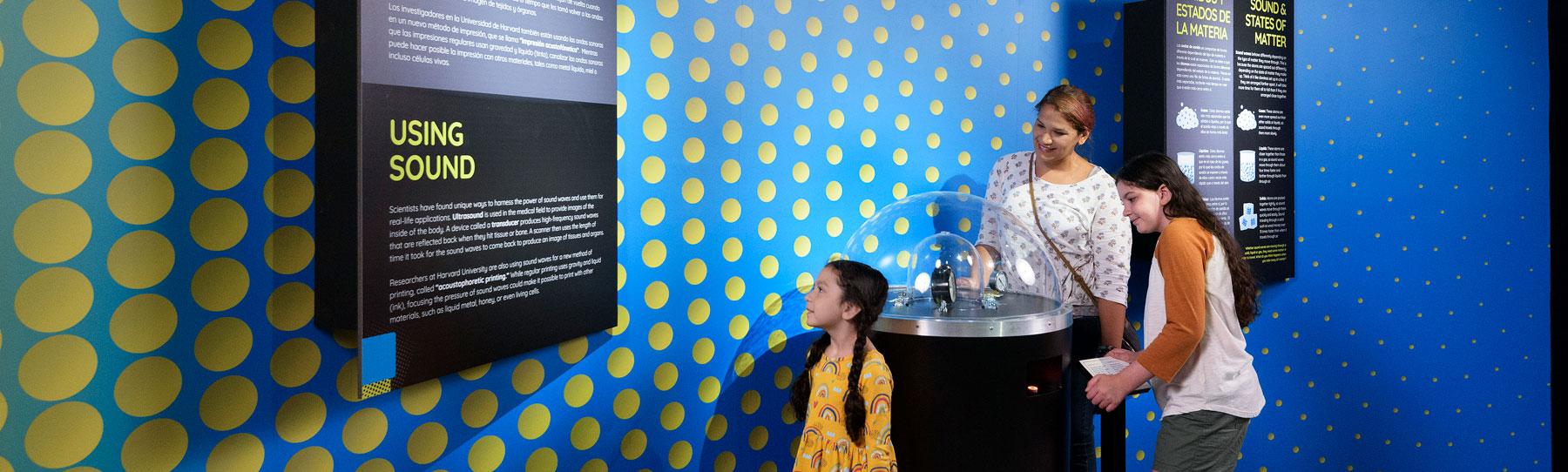 A mother and her two daughters look at hands-on demonstration and read a wall text panel in the Science of Sound exhibit.