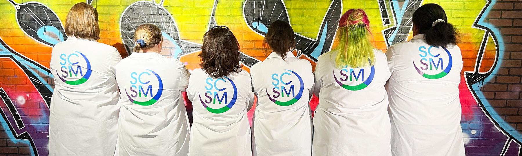 Group stand with backs to camera showing the SCSM logo on the backs of their lab coats