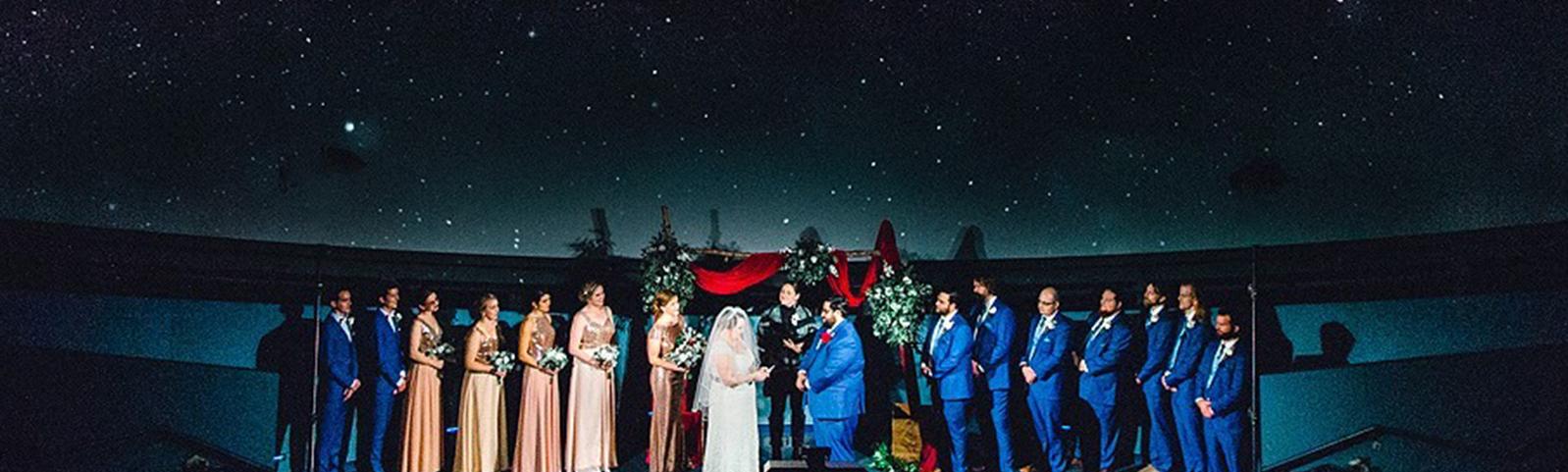 Wedding ceremony in the planetarium with couple flanked by bridesmaids and groomsmen with large projection of a starry night sky behind them.