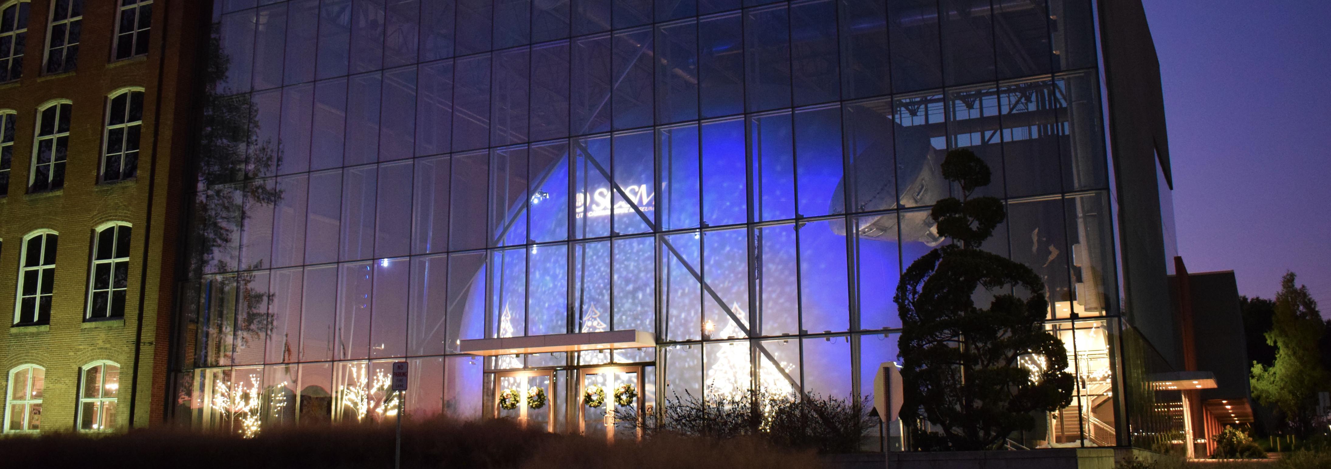 Four story building with glass walls with large dome inside. Dome is lit up in blue and white with snowflakes and the museum logo on the side.