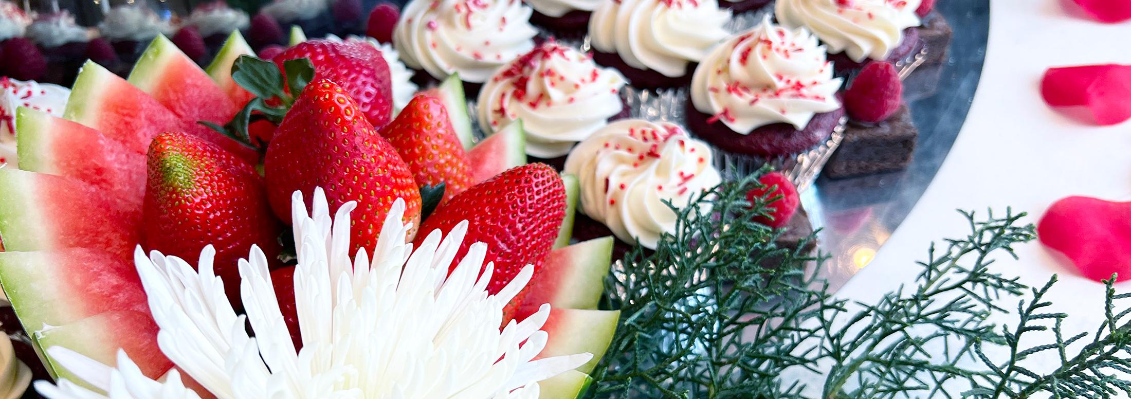 Slices of watermelon and Strawberries arranged to look like a flower with rows of cupcakes visible behind them and pink flower petals decorating the table.