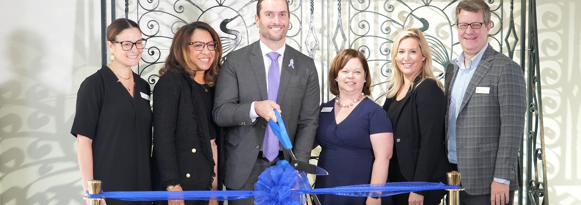 Group stands in front of a wrought iron gate on display with a blue ribbon in front of them. Man in center holds a giant pair of scissors as he prepares to cut the ribbon.