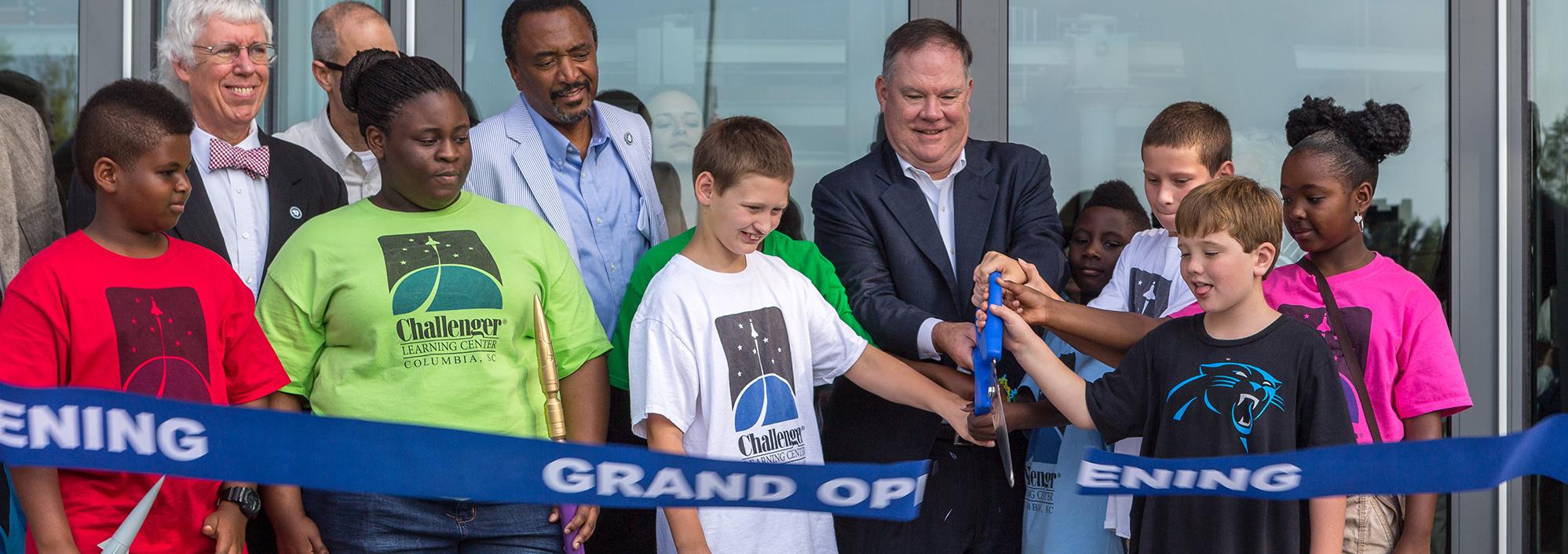 Group of older and younger people stand behind a blue ribbon that is being cut by a man holding an oversize pair of blue handled scissors.