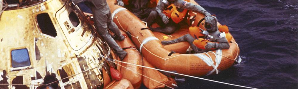 Space capsule and rescue raft shown in water. One individual is still on the capsule, three are in the raft.