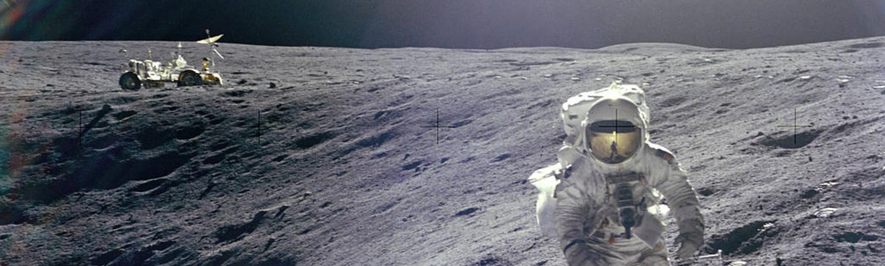 Astronaut Charles Duke stands on the Moon facing the camera next to a crater with the lunar rover at a distance in the background.