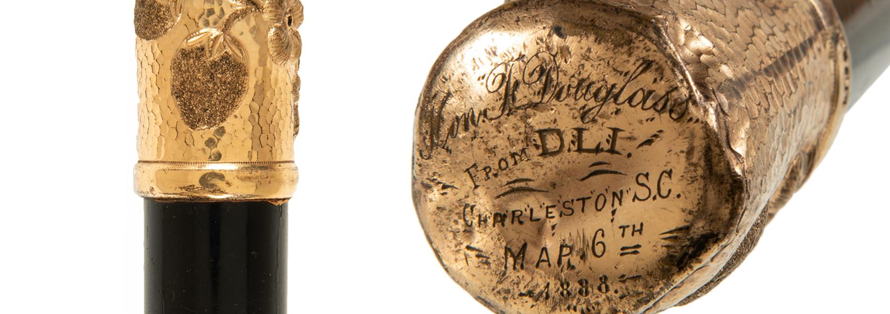 Showing the top of a walking stick. It is gold with fruit on the side and text on the top.