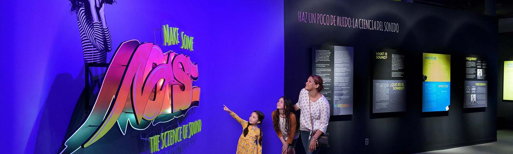 A Mother with two daughters look at the exhibit entrance which features a large sign with the exhibit title done in a graffiti art style.