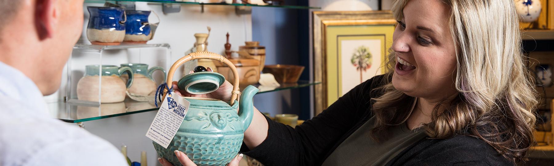 Blonde woman holds up light green ceramic tea pot on sale in the museum store