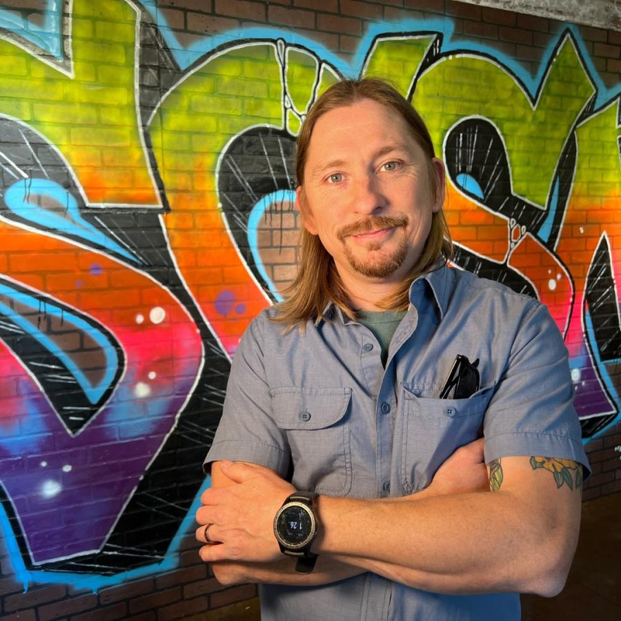 Man with shoulder length hair and mustache stands with arms crossed smiling at camera with wall in background that has a spray painted SCSM on it