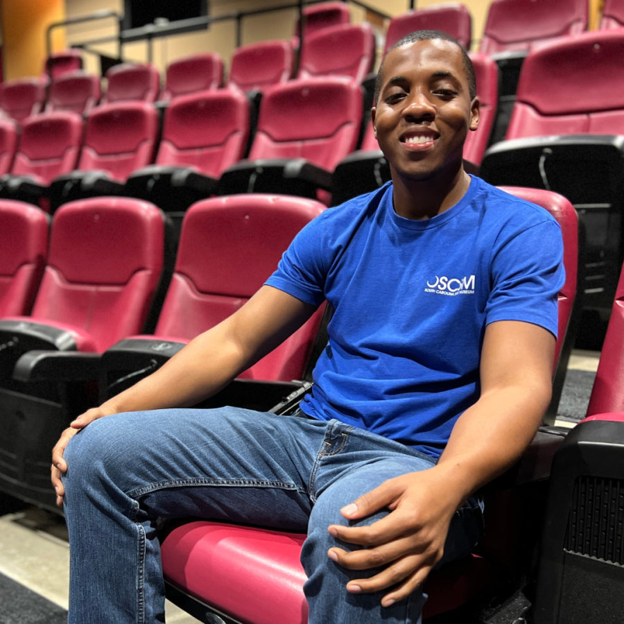Man in blue shirt with museum logo sits in red theater seat and smiles at camera