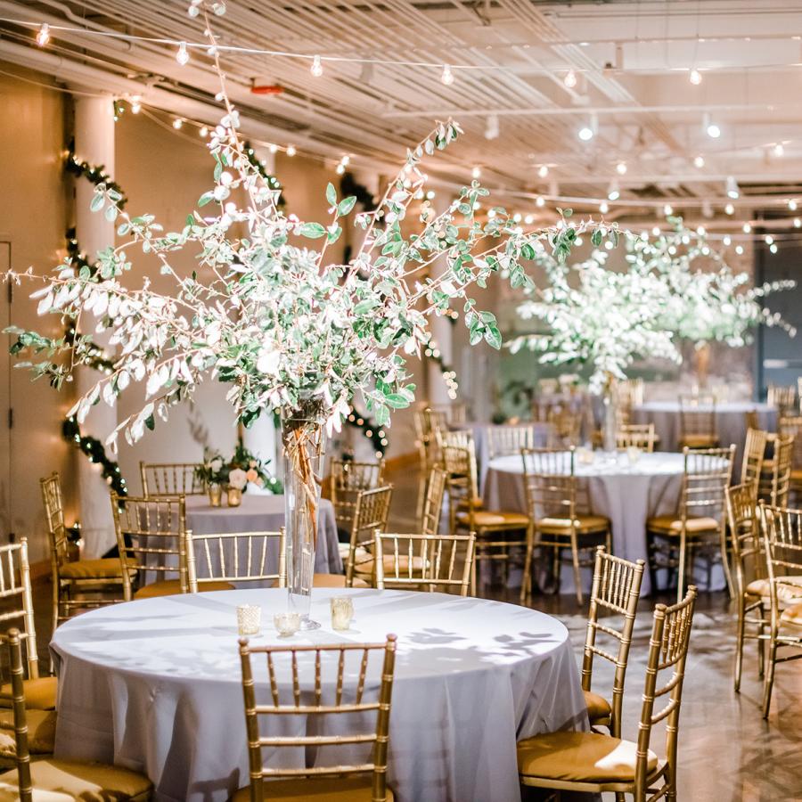 Large space with string lights suspended from ceiling and round tables with silver table cloths, gold chairs and large flower center pieces.