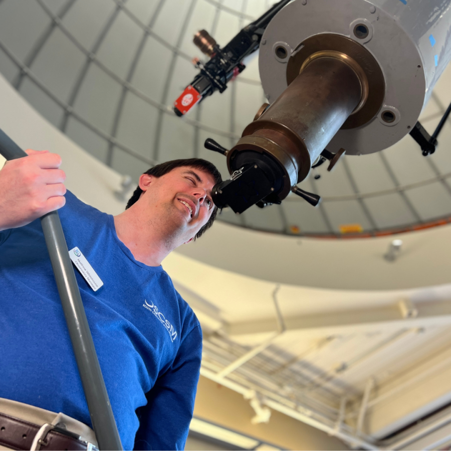 Man in long sleeved blue shirt looks through eyepiece of large white telescope