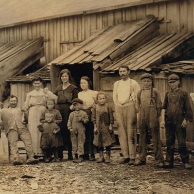 Old, sepia toned photograph depicts large group of adults and children in distressed clothing outside of a run down building