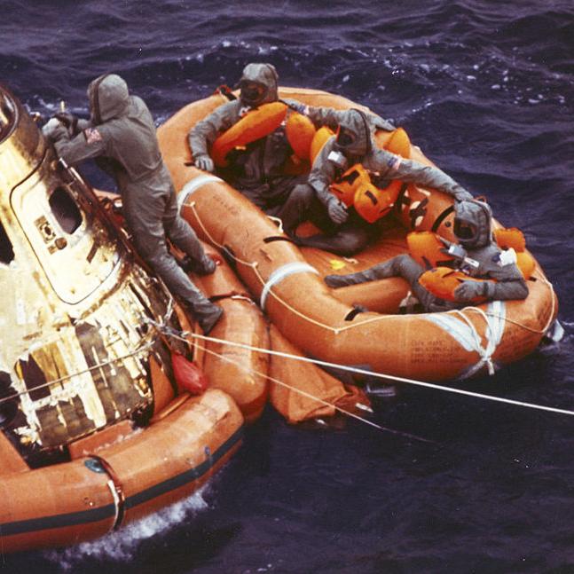 Space capsule and rescue raft shown in water. One individual is still on the capsule, three are in the raft.