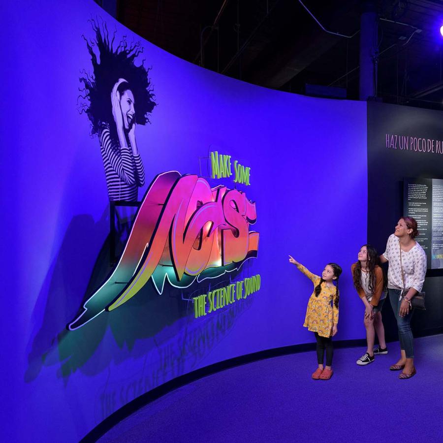A Mother with two daughters look at the exhibit entrance which features a large sign with the exhibit title done in a graffiti art style.