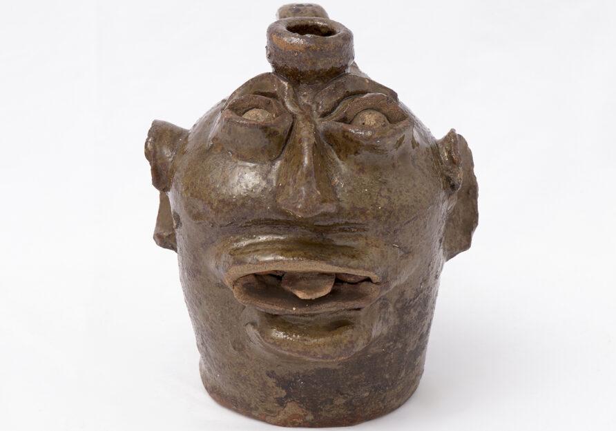 Small ceramic jug with odd looking face featuring large ears and nose, mouth open slightly with a protruding tongue.