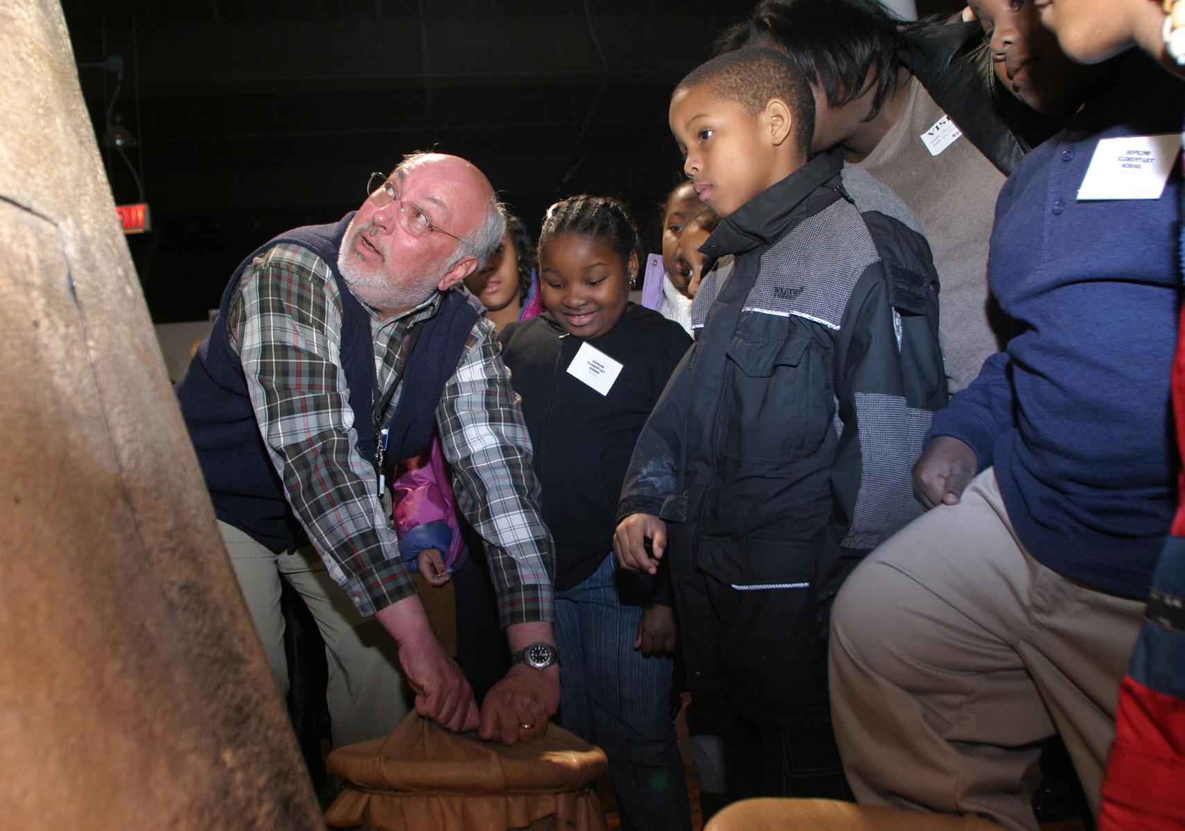 An older gentleman wearing a SCSM volunteer demonstrates an historic pot to a group of third grade students.