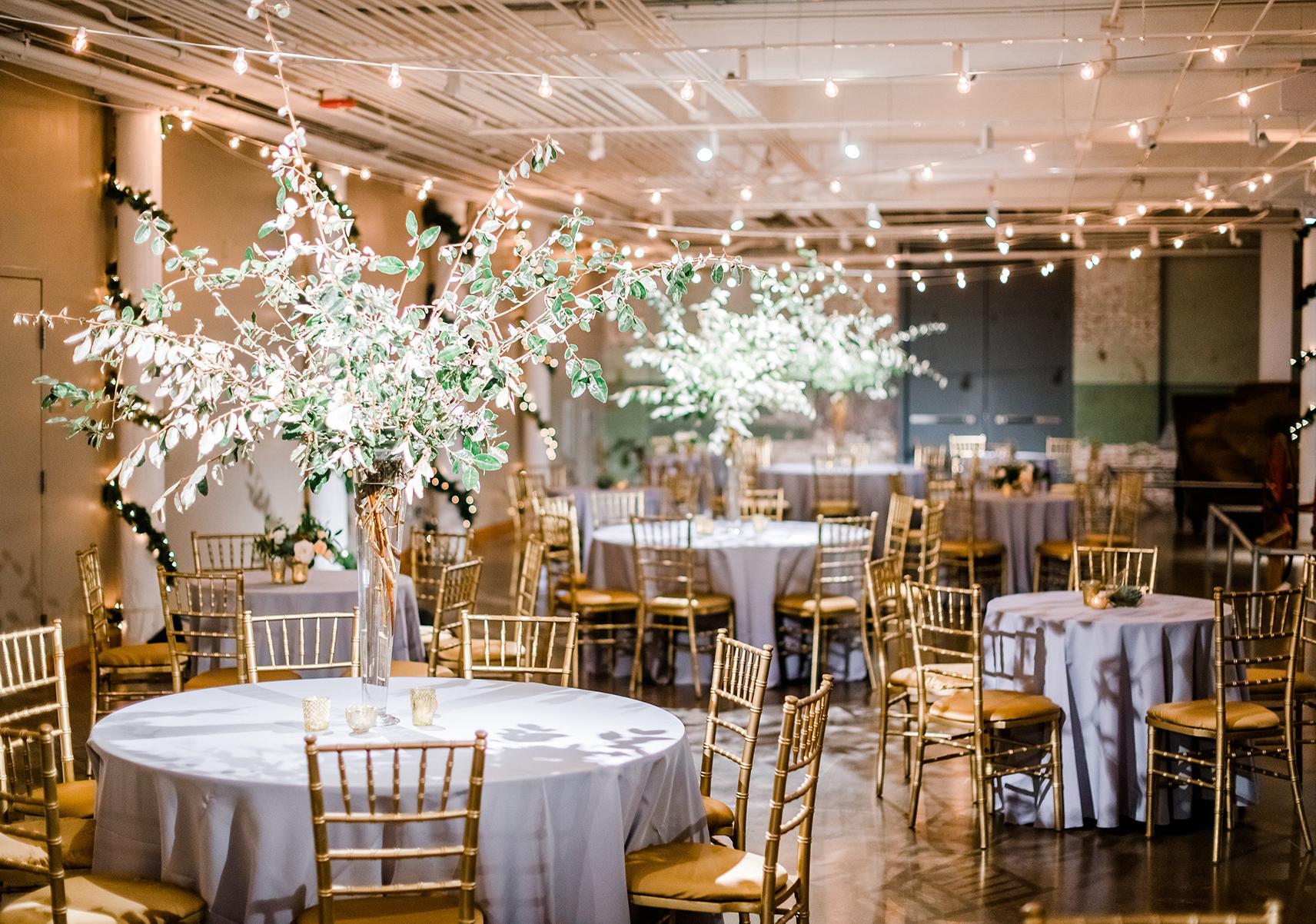 Large space with string lights suspended from ceiling and round tables with silver table cloths, gold chairs and large flower center pieces.