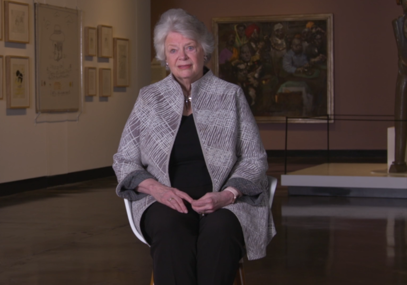 Woman in gray jacket and black top and pants sits in chair with artwork behind her.