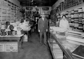 Black and white shows a man and woman standing in a shop with folded textiles in shelves on the walls.