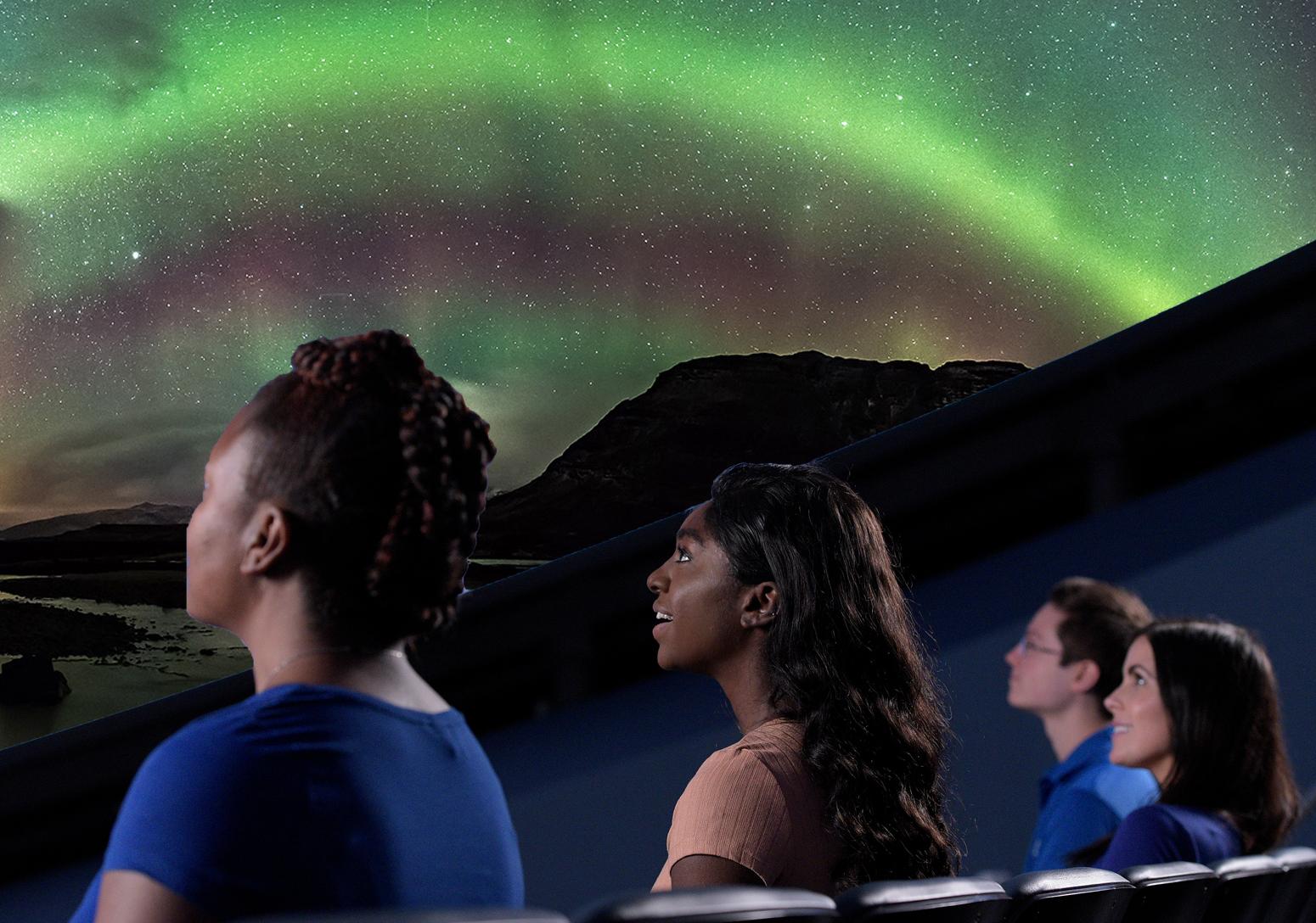 Three audience members sit in a row from left to right looking up at a screen showing a projection of bright glowing green lights