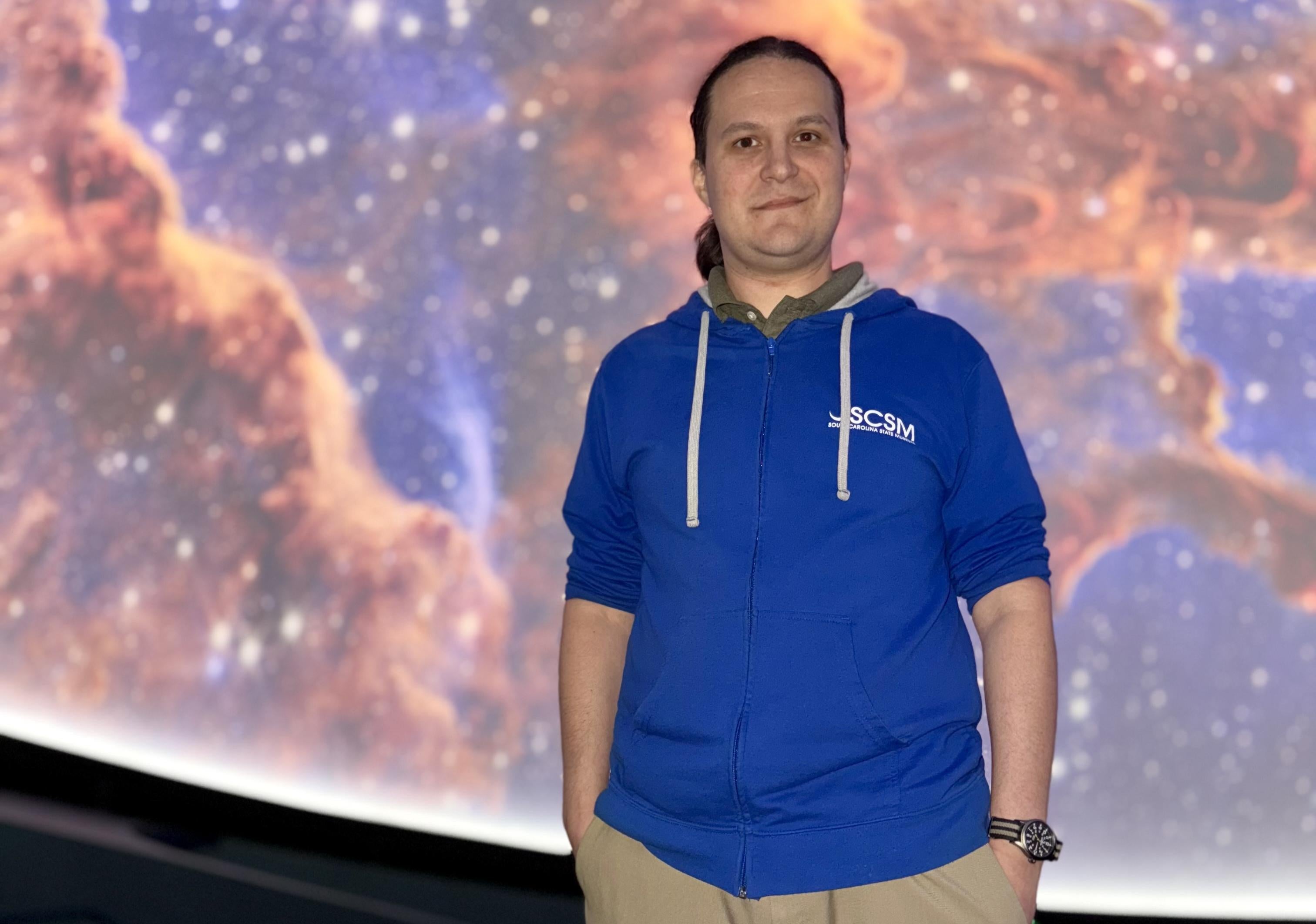 Man in blue hoodie and tan pants stands with a star nebula projected on a screen behind him.