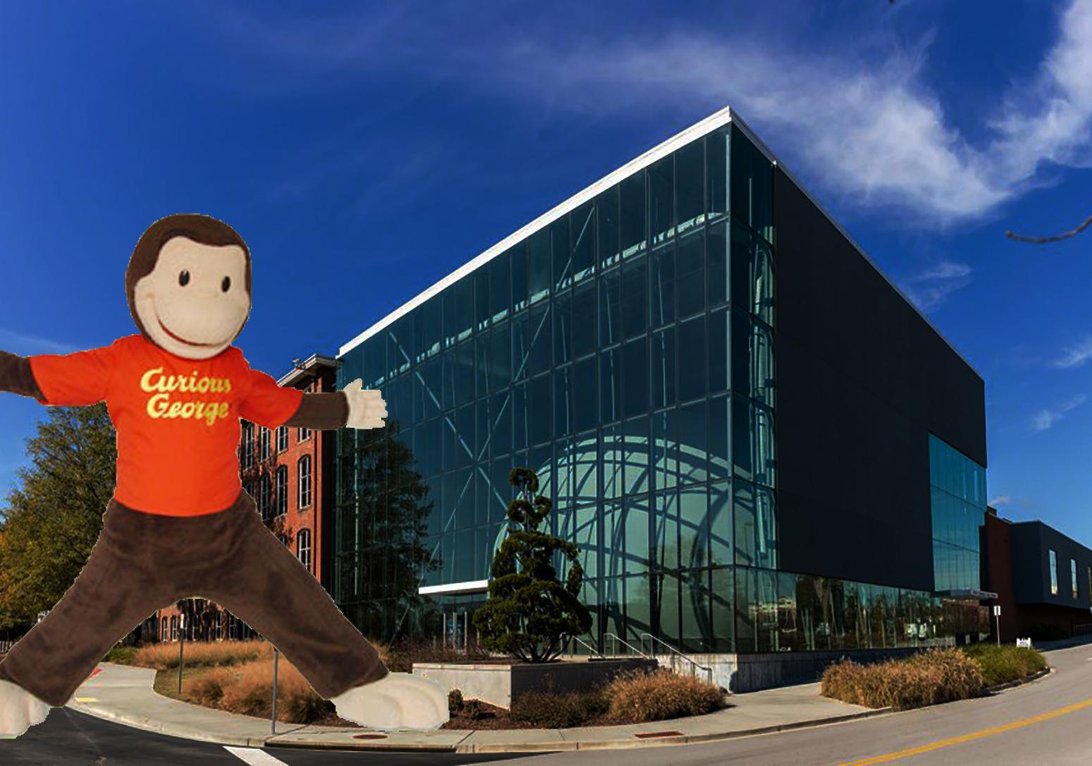 Curious George, a monkey stuffed animal with a red shirt with the outside of the South Carolina State Museum behind him