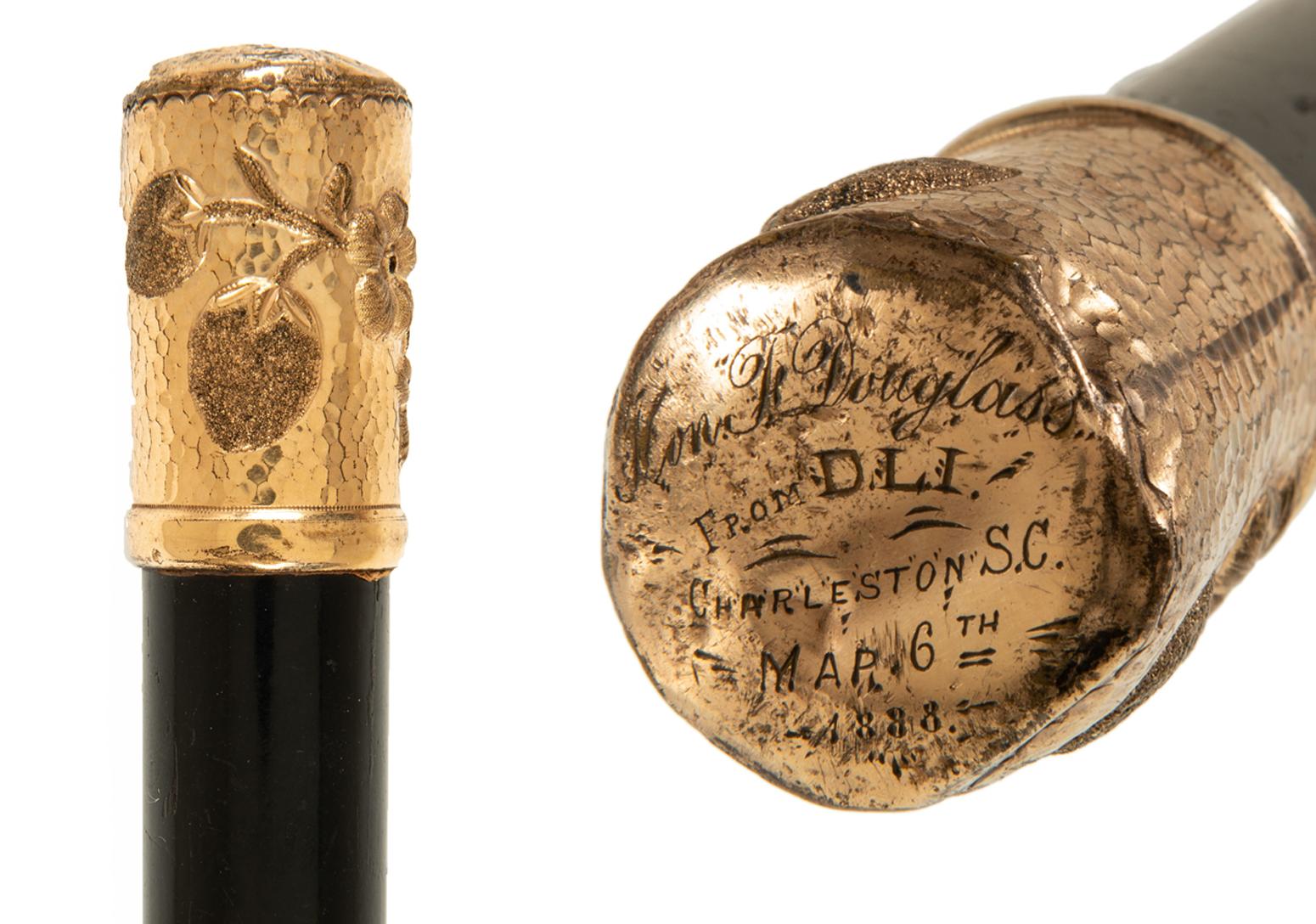 Showing the top of a walking stick. It is gold with fruit on the side and text on the top.