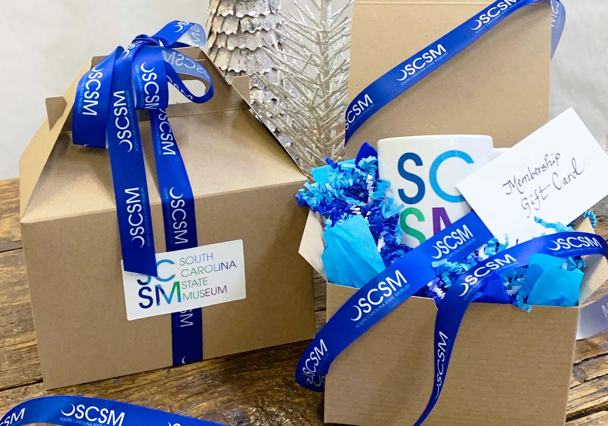 Two brown gift boxes on display with one partly open to show an SCSM mug inside with blue SCSM branded ribbon entwined